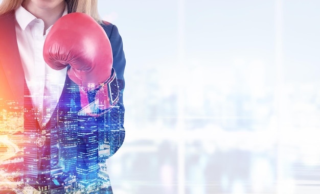 Gros plan d'une femme d'affaires blonde en costume portant des gants de boxe rouges et debout dans son bureau avec des fenêtres panoramiques. Paysage urbain. Maquette. Image tonique. Double exposition.