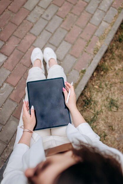 Gros plan d'une femme d'affaires en costume blanc et lunettes de soleil travaillant sur une tablette numérique assis près d'un immeuble de bureaux moderne dans un parc de la ville