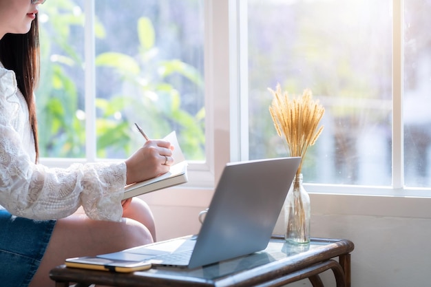 Gros plan d'une femme d'affaires indépendante asiatique prenant des notes écrites sur un ordinateur portable travaillant occasionnellement avec un ordinateur portable avec une tasse de café et un smartphone au caféBusiness Lifestyle