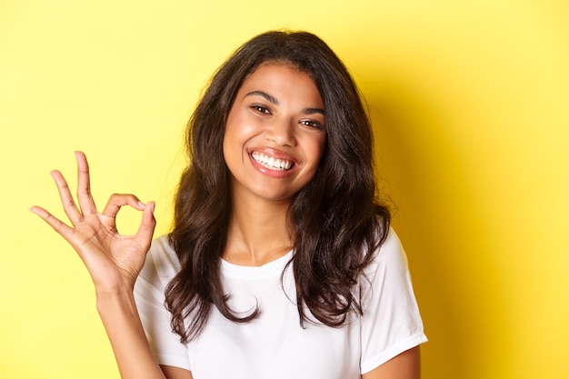 Gros plan d'une femme afro-américaine satisfaite, montrant un signe d'accord et souriant, recommandant quelque chose de bien, debout sur fond jaune.