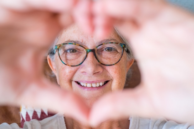 Gros plan d'une femme âgée faisant un geste en forme de coeur avec les mains. Femme âgée heureuse à la retraite faisant un symbole en forme de coeur avec ses deux mains et regardant la caméra