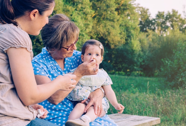 Gros plan d'une femme âgée se nourrissant de purée de fruits à une adorable petite fille assise sur un banc à l'extérieur. Concept de trois générations féminines différentes.