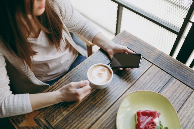 Gros plan sur une femme assise dans un café avec une tasse de cappuccino, un gâteau, se relaxant au restaurant pendant le temps libre. Femme tenant un téléphone portable avec un écran vide vide, reste au café. Concept de mode de vie