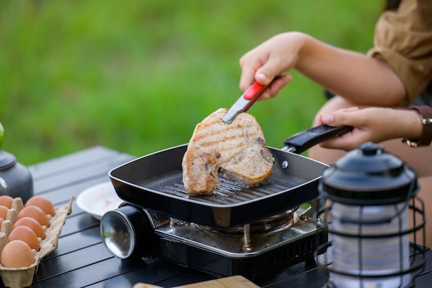 Gros plan d'une femme au barbecue de porc grillé dans la poêle à pique-nique et la cuisson des aliments tout en étant assis sur une chaise dans le camping