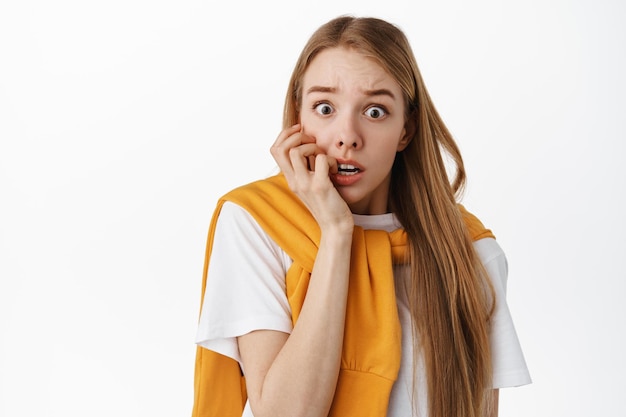 Gros plan d'une femme blonde choquée et effrayée, mordant les doigts et haletant, regardant surpris et effrayé, effrayée par quelque chose d'effrayant, debout sur fond blanc