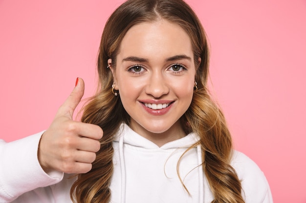 Gros plan femme blonde souriante portant des vêtements décontractés montrant le pouce vers le haut et regardant l'avant sur le mur rose