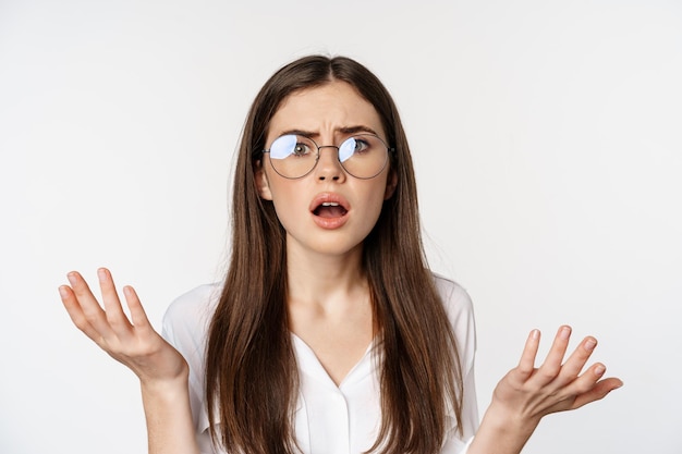 Gros plan d'une femme brune confuse dans des verres qui a l'air perplexe et désemparée debout sur le dos blanc...