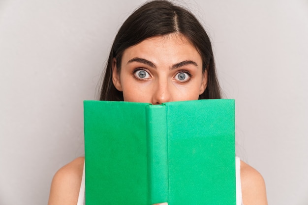 gros plan d'une femme brune heureuse portant une robe et couvrant son visage avec un livre isolé sur un mur gris