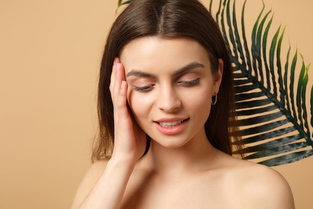 Gros plan sur une femme brune à moitié nue avec une peau parfaite, un maquillage nu et une feuille de palmier isolée sur un mur pastel beige