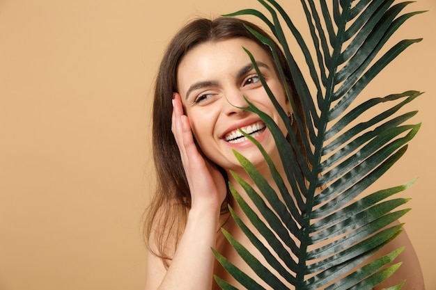 Gros plan sur une femme brune à moitié nue avec une peau parfaite, un maquillage nu et une feuille de palmier isolée sur un mur pastel beige