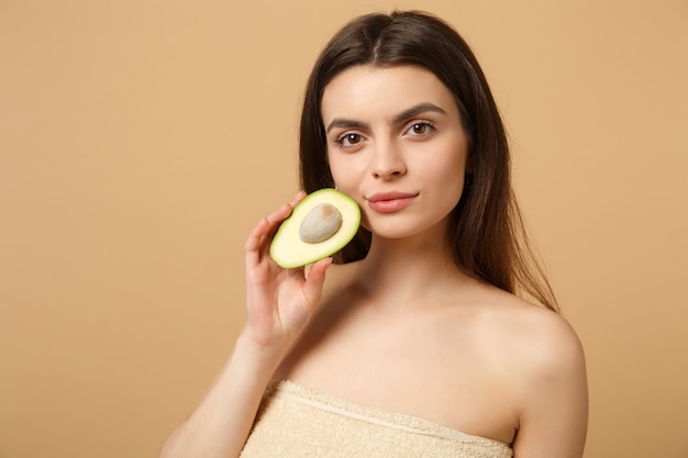 Gros plan sur une femme brune à moitié nue avec une peau parfaite, un maquillage nu tenant un avocat isolé sur un mur pastel beige