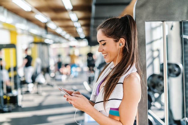 Gros plan d'une femme caucasienne souriante, choisir la musique sur un téléphone intelligent en se tenant debout dans une salle de sport. Serviette autour du cou.