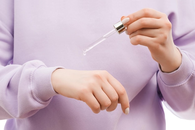 Gros plan d'une femme dans un sweat-shirt surdimensionné lavande tenant une pipette avec du sérum dans les mains La goutte de sérum est suspendue De beaux ongles soignés avec du vernis à ongles