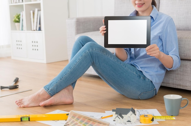 gros plan d'une femme designer d'intérieur de maison souriante montrant un écran vide avec une tablette numérique mobile et assise dans le plancher en bois du salon faisant du travail.