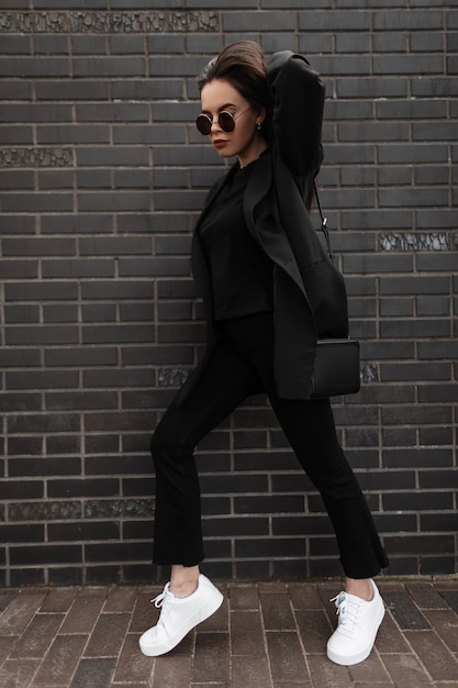 Gros plan femme élégante en mode veste longue en pantalon denim vintage en t-shirt avec sac à main en cuir noir à la mode près d'un bâtiment en briques dans la rue. Tenue de printemps tendance. Mode casual.