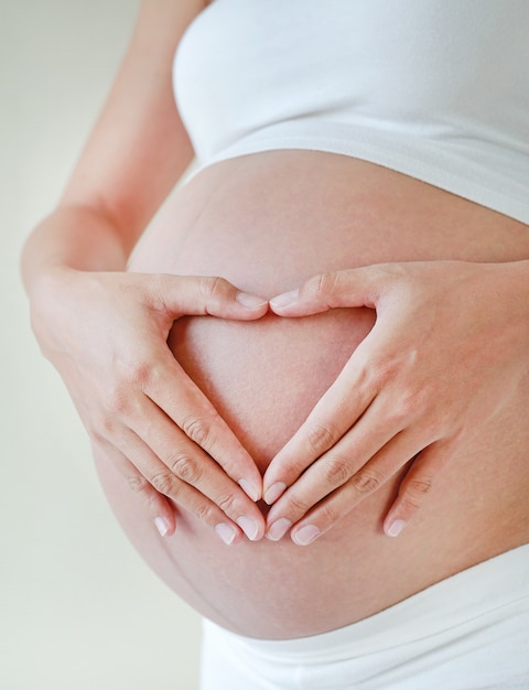 Gros plan femme enceinte les mains en forme de coeur sur son ventre.