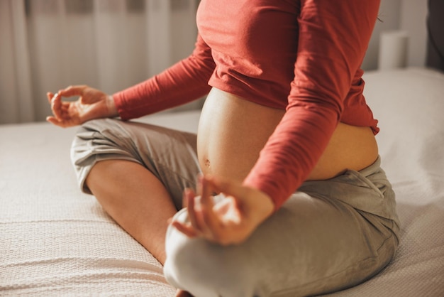 Gros plan d'une femme enceinte méconnaissable faisant des exercices de méditation de yoga en position du lotus sur un lit dans la chambre.