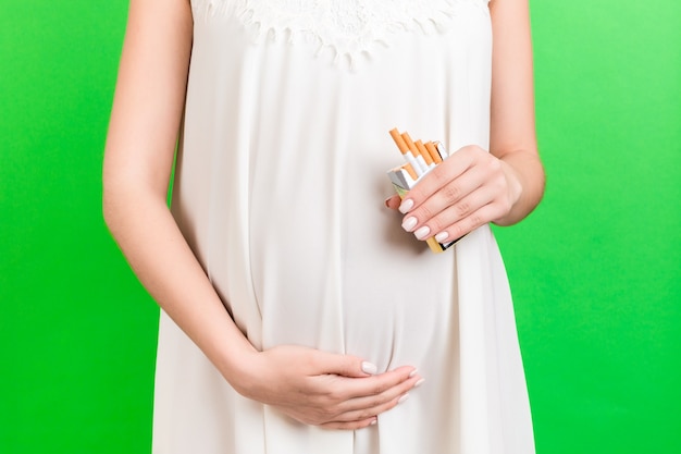 Gros plan sur une femme enceinte en robe blanche fumant une cigarette sur fond vert. Abus de nicotine et de tabac. Habitude dangereuse pour un futur bébé.