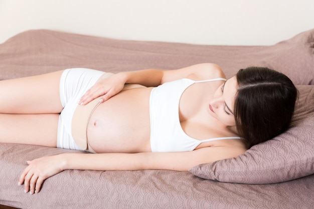 Gros plan femme enceinte robes bandage sur le ventre à la maison sur le lit ceinture de soutien abdominale orthopédique