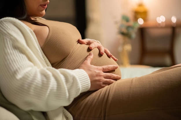 Gros plan d'une femme enceinte tenant le ventre. Femme en robe et cardigan assis sur le lit. Grossesse, concept d'attente