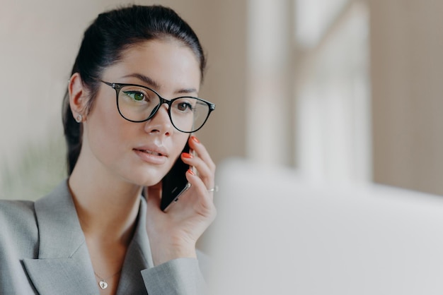 Gros plan d'une femme européenne brune à l'air agréable porte des lunettes transparentes concentrées sur l'écran d'un ordinateur portable a une conversation téléphonique pendant le travail à distance travaille sur le concept de technologie de projet