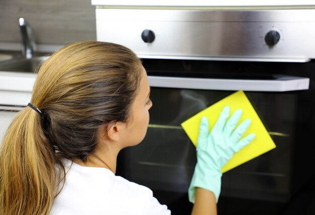 Gros plan d'une femme avec des gants de protection pour nettoyer la porte du four. Fille de polissage de cuisine. Gens, travaux ménagers, concept de nettoyage.