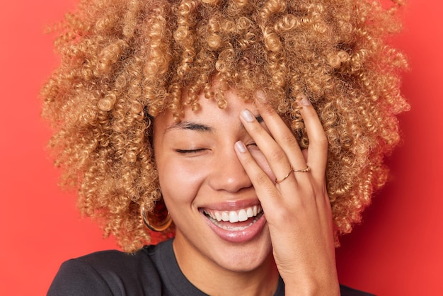 Gros plan d'une femme heureuse et insouciante fait face à la paume garde les yeux fermés sourit à pleines dents montre des dents blanches porte des boucles d'oreilles t-shirt noir isolé sur fond rouge Concept d'émotions positives