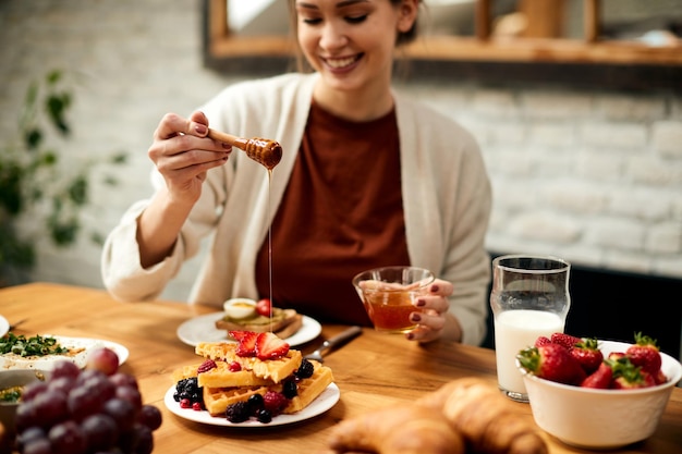 Gros plan d'une femme heureuse versant du miel sur des gaufres pendant le petit-déjeuner à la table à manger