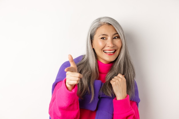 Gros plan d'une femme hipster senior heureuse et cool, vérifiant, pointant et regardant la personne, souriant sur le côté, debout sur fond blanc.