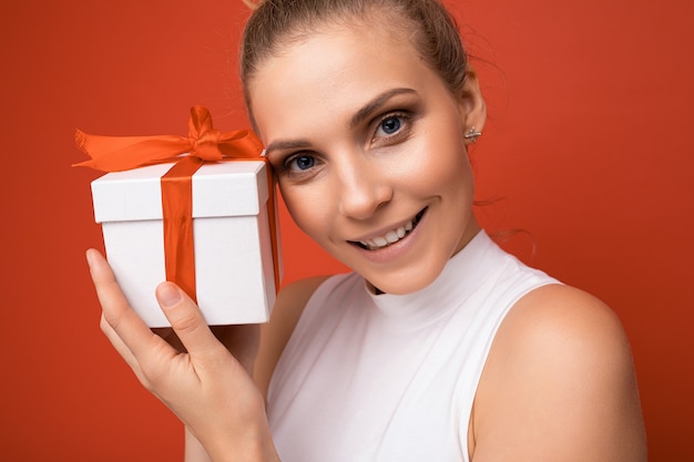 Gros plan d'une femme isolée sur un mur de fond coloré portant une tenue tendance tous les jours tenant une boîte-cadeau et regardant la caméra.