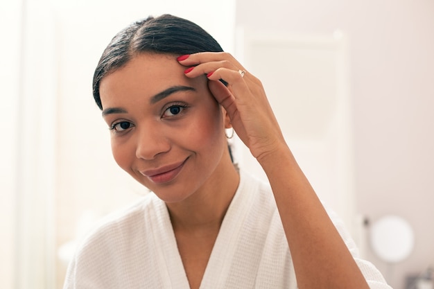 Gros plan de la femme joyeuse et calme debout en peignoir et souriant tout en touchant les cheveux