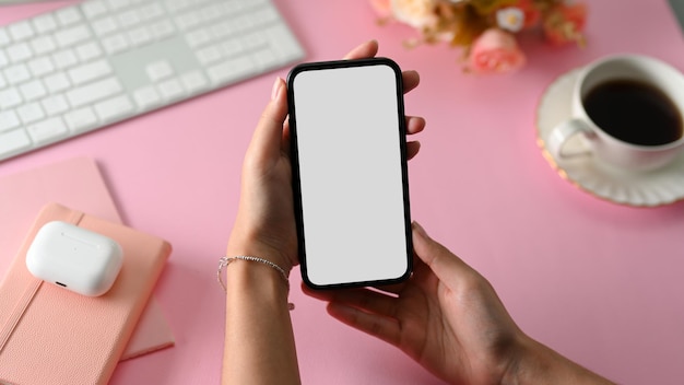 Gros plan Femme main tenant une maquette d'écran blanc de téléphone sur une table d'espace de travail de bureau rose féminin