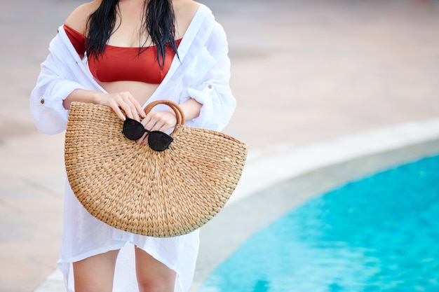 Gros plan d'une femme méconnaissable en chemise de plage blanche debout avec un sac de paille et des lunettes de soleil au bord de la piscine