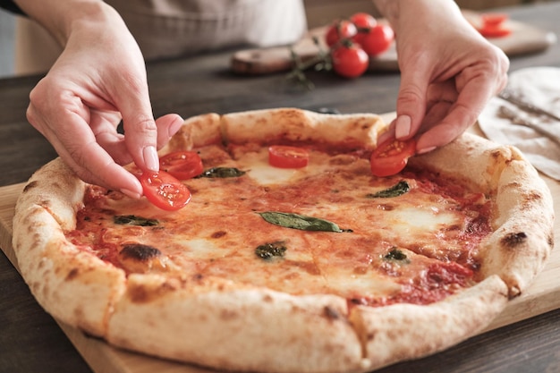 Gros plan d'une femme méconnaissable décorant une pizza au fromage avec des tranches de tomate après la cuisson