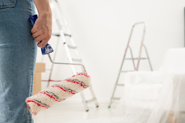 Gros plan d'une femme méconnaissable en jeans tenant le rouleau de peinture dans la chambre avec escabeaux