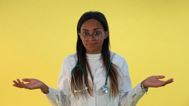 Photo gros plan de femme médecin de race mixte, étalant les mains sur les côtés sur fond jaune.