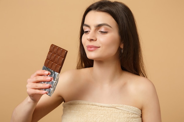 Gros plan sur une femme à moitié nue avec une peau parfaite, un maquillage nu tient une barre de chocolat isolée sur un mur pastel beige