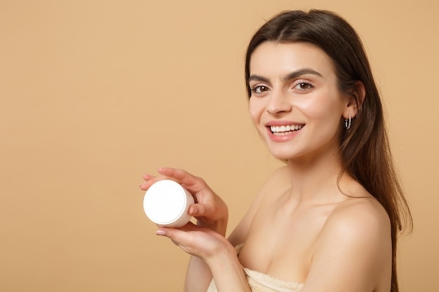 Gros plan femme à moitié nue avec une peau parfaite maquillage nude appliquant une crème pour le visage isolée sur un mur pastel beige