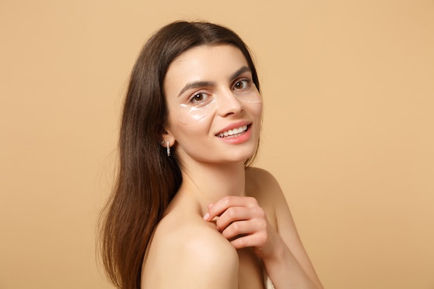 Gros plan sur une femme à moitié nue avec une peau parfaite, des patchs de maquillage nus sous les yeux isolés sur un mur pastel beige