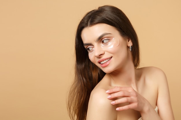 Gros plan sur une femme à moitié nue avec une peau parfaite, des patchs de maquillage nus sous les yeux isolés sur un mur pastel beige