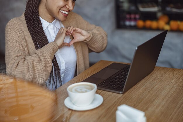 Gros plan d'une femme multiethnique tenant les doigts en forme de coeur et parlant par appel vidéo sur un ordinateur portable dans un café