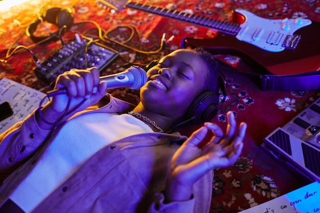 Gros plan d'une femme noire souriante tenant un microphone et chantant allongé sur un tapis dans un néon violet