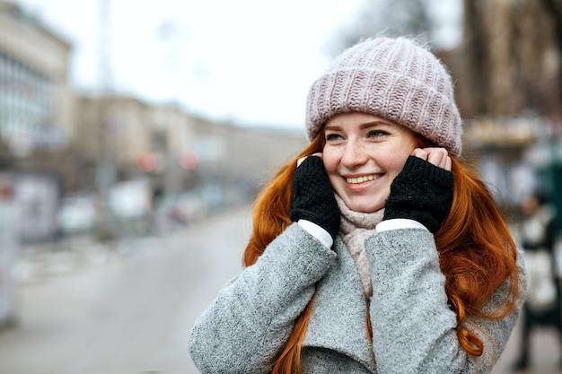 Gros plan d'une femme romantique au gingembre aux cheveux longs portant un bonnet et une écharpe tricotés marchant dans la ville. Espace pour le texte