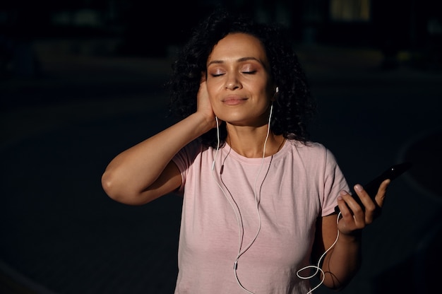 Gros plan sur une femme sereine avec des écouteurs, tenant un téléphone portable et appréciant d'écouter de la musique les yeux fermés