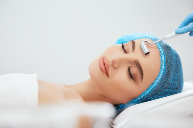 Gros plan d'une femme souriante au bonnet bleu allongé sur un canapé dans une clinique cosmétologique avec les yeux fermés. Les mains du médecin dans des gants bleus faisant la procédure avec électrode pour la galvanisation