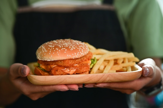 Gros plan femme tenant un savoureux hamburger de poisson fait maison avec un plateau en bois de pommes de terre frites.