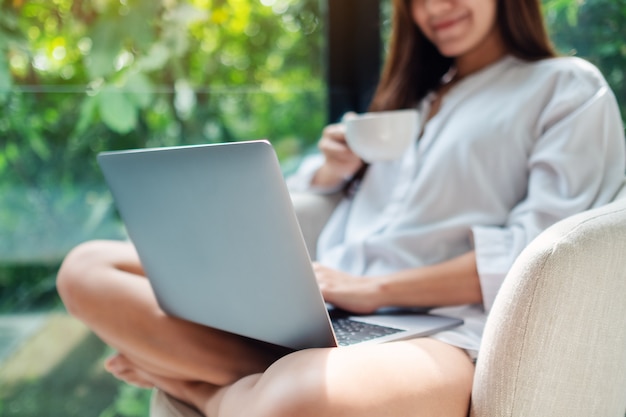 Gros plan d'une femme travaillant sur un ordinateur portable tout en buvant du café et assis sur un fauteuil blanc à la maison avec la nature verte