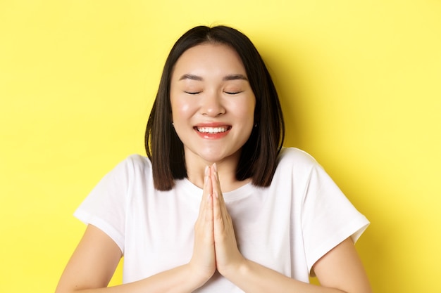 Gros plan d'une fille asiatique pleine d'espoir en t-shirt blanc, main dans la main en priant, geste namste et souriant, faisant un souhait ou plaidant, debout sur le jaune