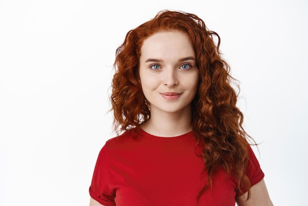 Gros plan d'une fille au gingembre avec des cheveux bouclés et un maquillage de lumière naturelle souriant à la caméra et regardant mignon debout en t-shirt rouge sur fond blanc