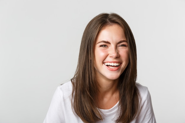 Gros plan d'une fille brune heureuse et insouciante souriant et riant de joie, debout blanc.